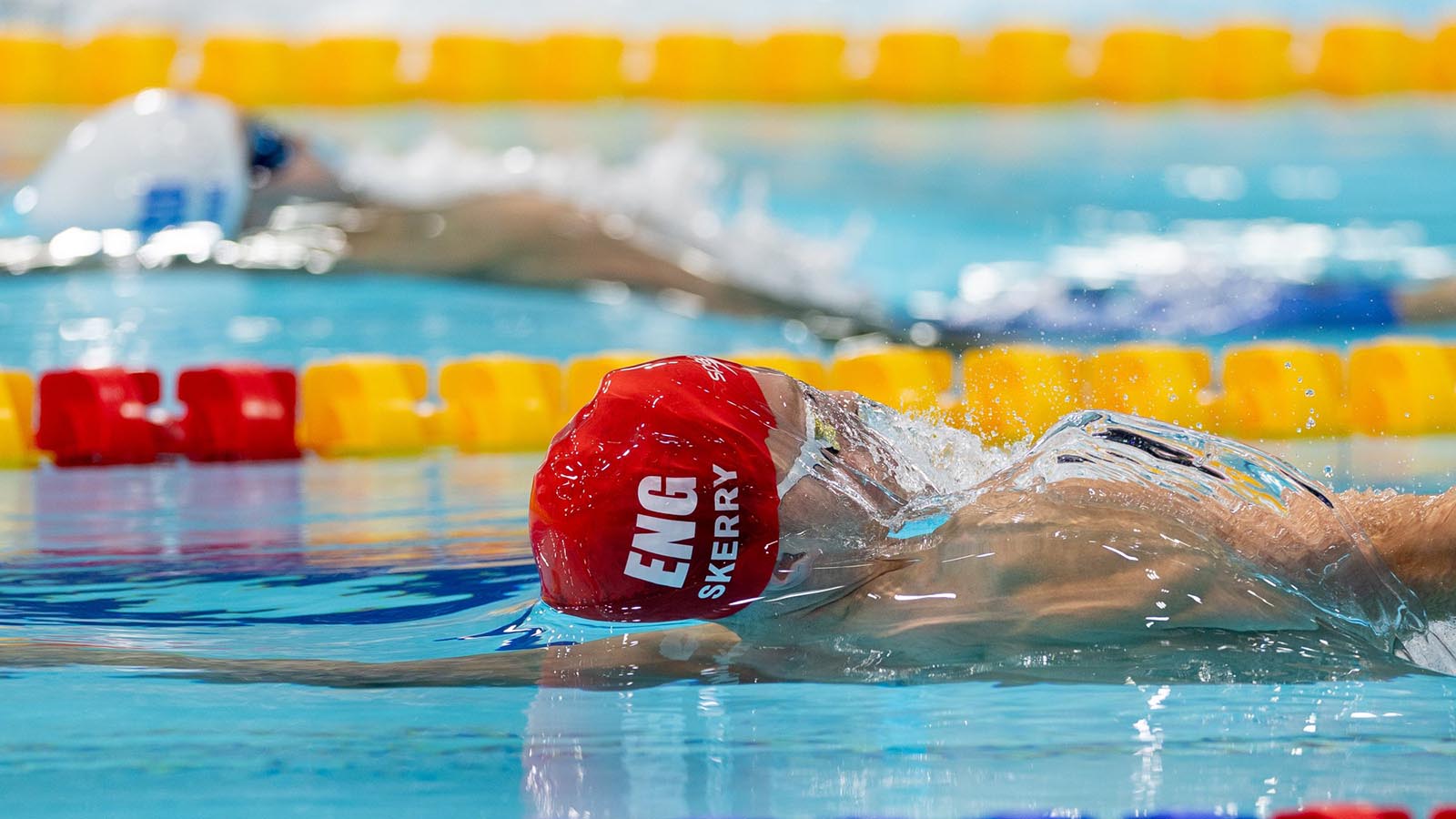 Two swimmers doing the backstroke in a race, one wearing a red cap and the other in a white cap