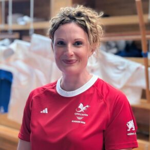 A portrait picture of Team Bath physio Katy Williams, wearing the Team Wales Glasgow 2026 Commonwealth Games kit, in front of some judogi hanging on the wall of a dojo.