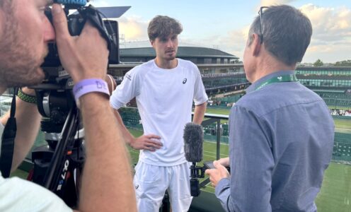 A film camera points at Team Bath Tennis player Toby Samuel as he is interviewed by BBC Points West sports reporter Ali Durden with Wimbledon Centre Court in the background.