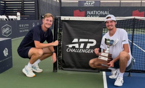 Coach Tiarnan Brady (left) and tennis player Toby Samuel kneeling next to a tennis net featuring the ATP Challenger Tour logo. Toby is holding a trophy.