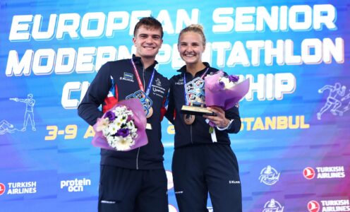 Pentathlon GB athletes Ross Charlton and Emma Whitaker, both wearing GB tracksuits, holding medals and flowers in front of a display screen showing the words "European Senior Modern Pentathlon Championship"