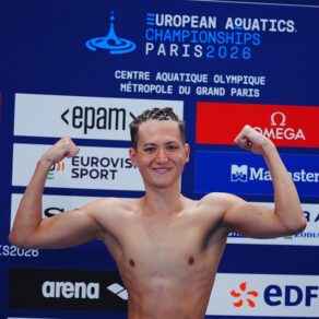 Artistic swimmer Ranjuo Tomblin flexing his muscles in front of a 2026 European Aquatics Championships sponsors board