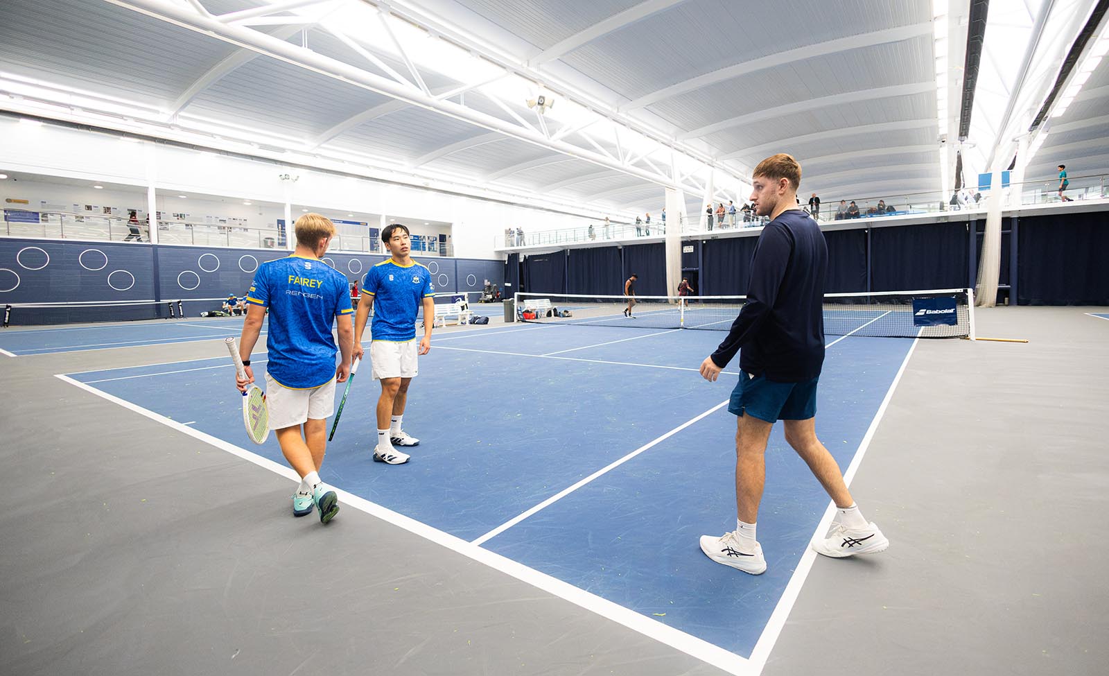 A tennis coach talks to two players on an indoor court
